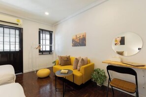A modern living room with a sofa, a round table, and a chair. There is a window with blinds, a wall-mounted mirror, and a picture frame. - Ombra House No26 (Chiang Mai)