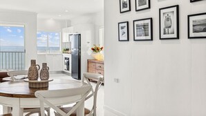 A modern kitchen with white cabinets, a dining area with a round table and chairs, and a view of the sea through large windows. - Boorolong 4 - Affordable Beachfront Escape with Convent Beach Views - Holiday Yamba (Yamba)