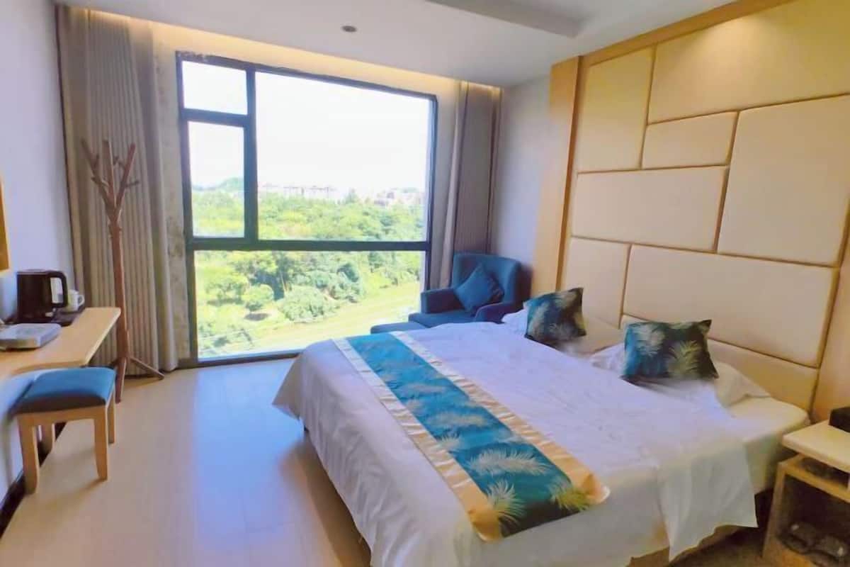 Quarto de hotel com uma cama grande, uma escrivaninha, uma cadeira e vista para o verde através de uma janela.