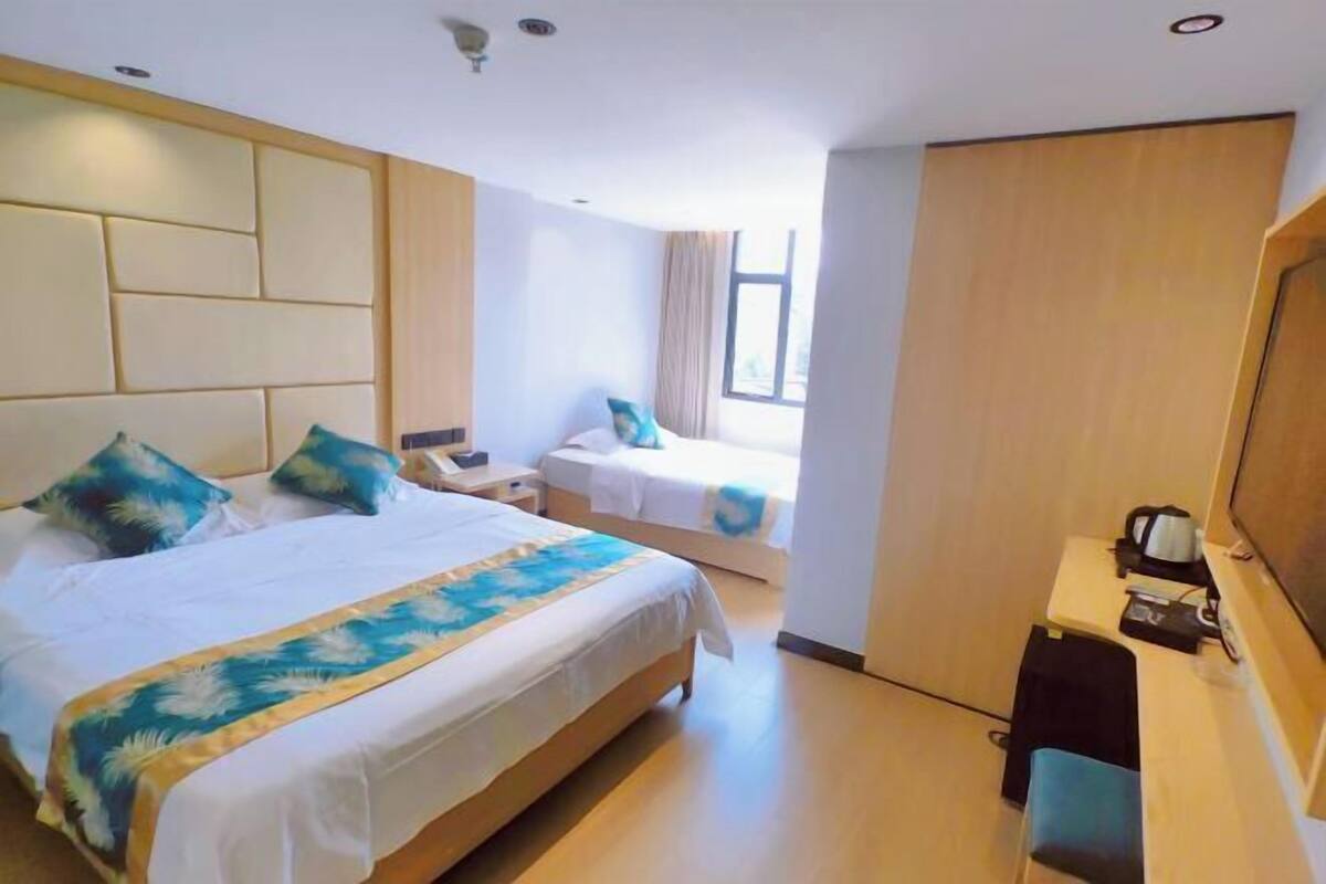 Quarto de hotel com uma cama grande, uma cama menor, uma escrivaninha com cadeira, televisão e janela com cortinas.