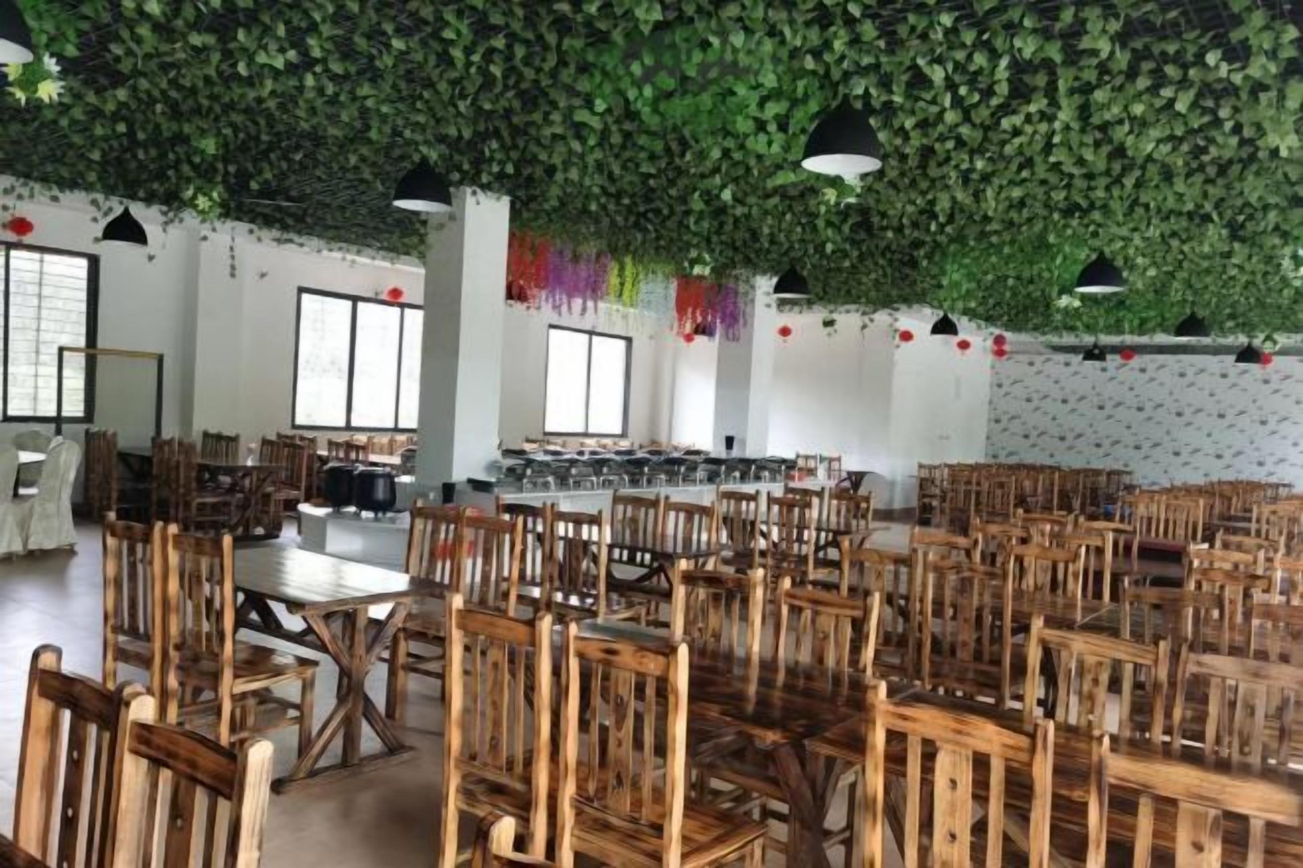 A spacious dining area with wooden tables and chairs, large windows, and a ceiling covered in green foliage.