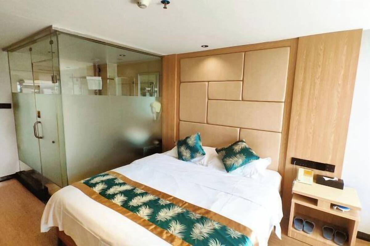 Quarto de hotel com uma cama grande, box de vidro e cabeceira de madeira.