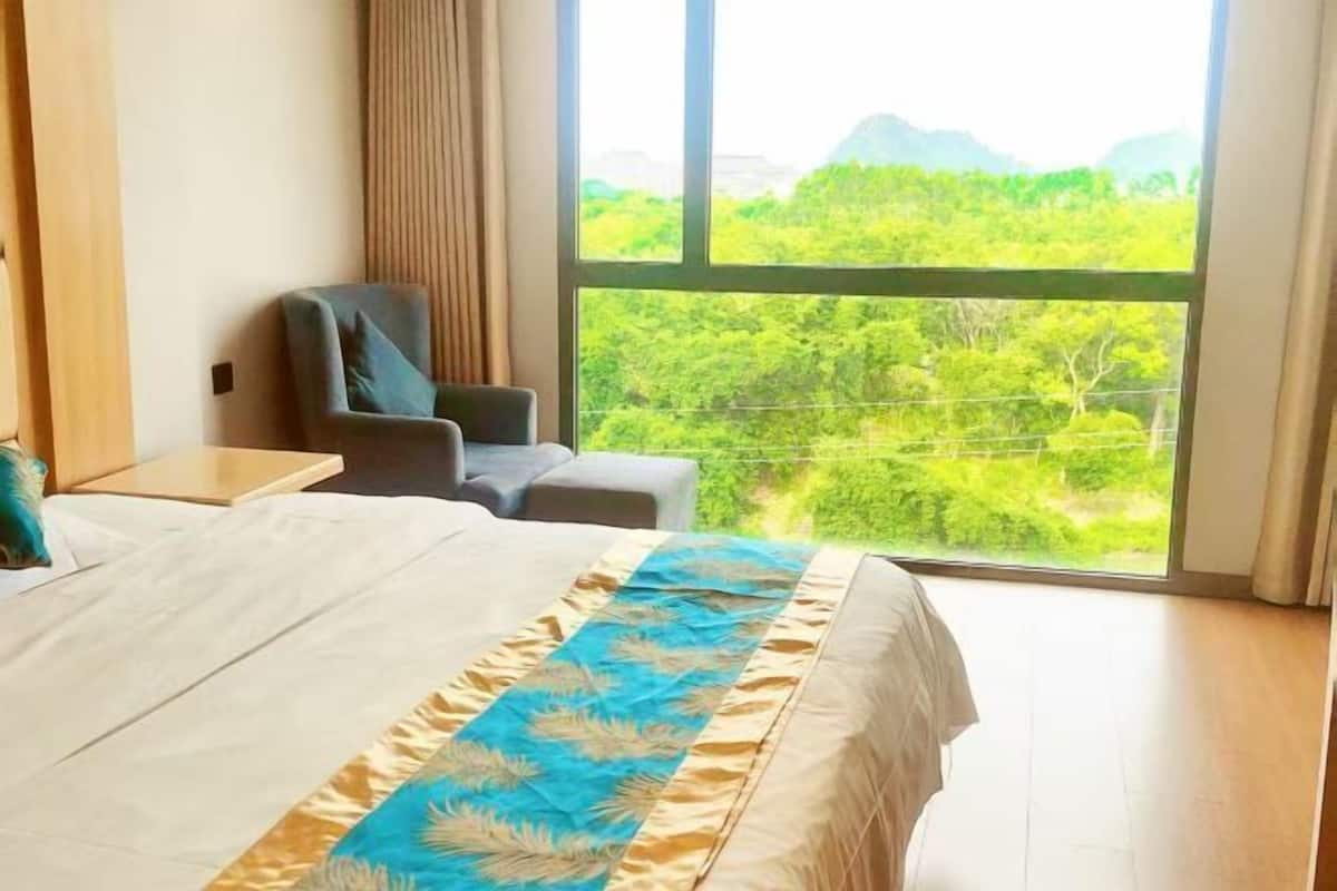 Quarto de hotel com uma cama grande, uma cadeira, uma mesinha e uma janela com vista para o verde.