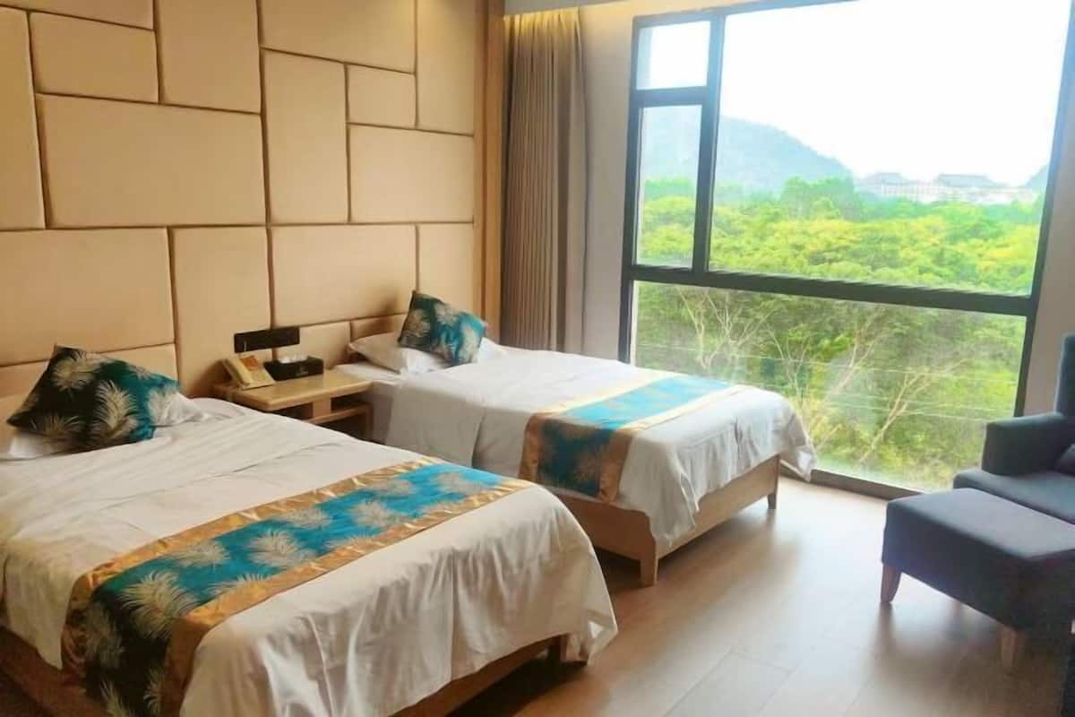 Quarto de hotel com duas camas, uma escrivaninha e uma cadeira. Há uma janela grande com vista para áreas verdes e colinas.