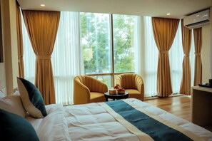 Habitación de hotel con una cama grande, dos sillones, una mesita y vistas a zonas verdes por las ventanas.