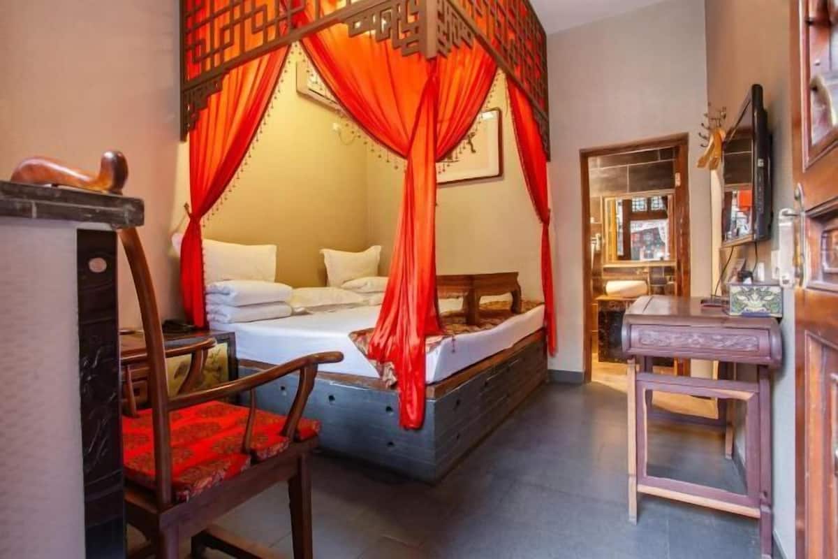 Ein traditionell eingerichtetes Zimmer mit einem Bett, einem Holzsessel, einem kleinen Tisch und einem Spiegel.
