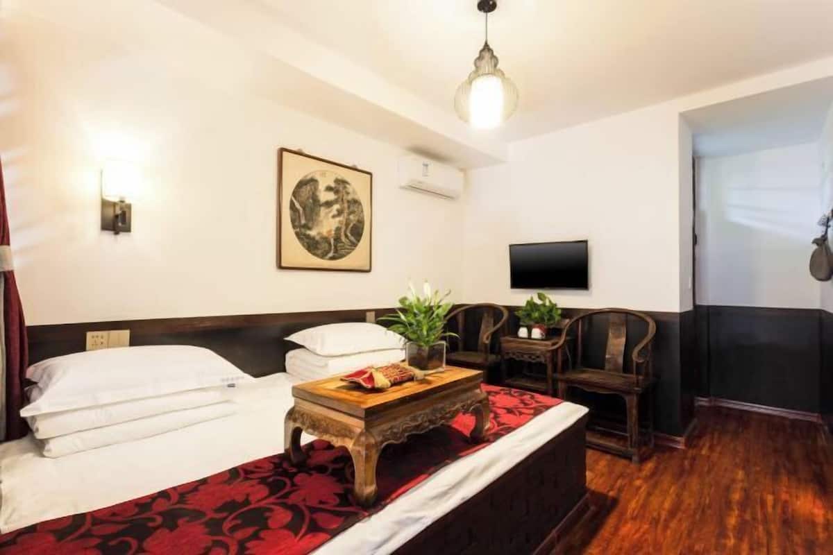 Ein Hotelzimmer mit einem Bett, einem kleinen Tisch mit Pflanze, einem an der Wand montierten Fernseher und einem Gemälde.