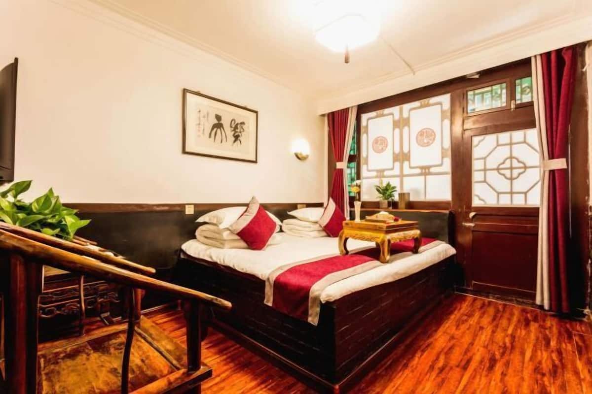 Ein traditionell eingerichteter Raum mit einem hölzernen Bett, einem kleinen Tisch mit Tablett, einer Wandleuchte und einem Fenster mit dekorativem Sichtschutz.
