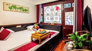 Ett hotellrum med en säng, ett litet träbord med en vas och ett bricka, en tv och ett fönster med dekorativa glasrutor.