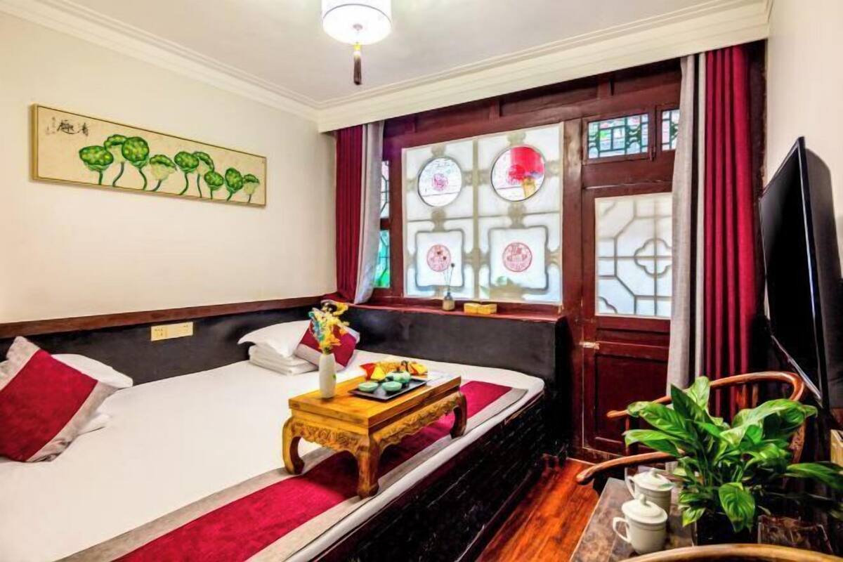 Ein Hotelzimmer mit einem Bett, einem kleinen Holztisch mit einer Vase und einem Tablett, einem Fernseher und einem Fenster mit verzierten Glasscheiben.