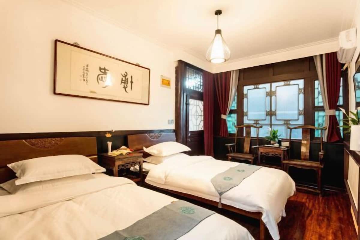 Ein Hotelzimmer mit zwei Betten, einem hölzernen Kopfteil, einem gerahmten Kunstwerk an der Wand, einem Schreibtisch mit Stuhl und einem Fenster mit Vorhängen.