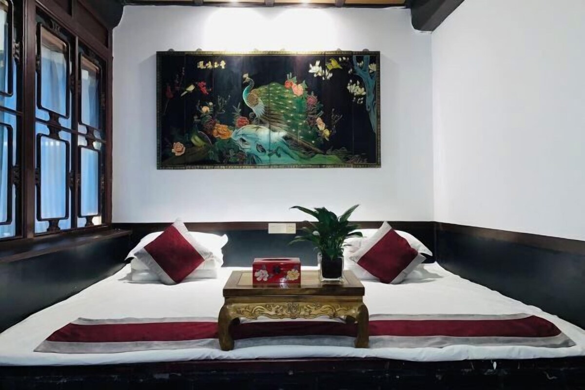 A hotel room with a bed, a decorative painting, a small table with a plant, and a window with curtains.