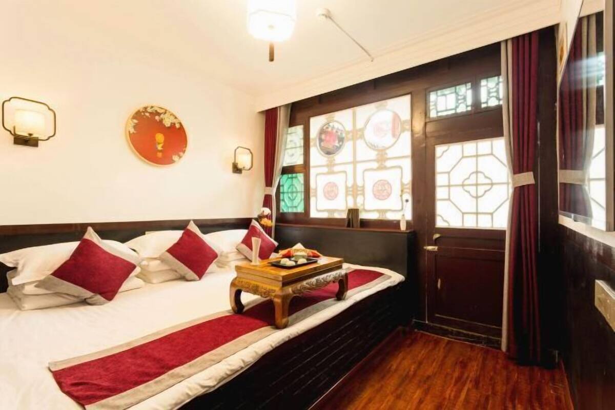 Ein Bett mit roter und weißer Bettwäsche, ein Nachttisch aus Holz mit einer Ablage und ein Fenster mit geometrischem Muster.
