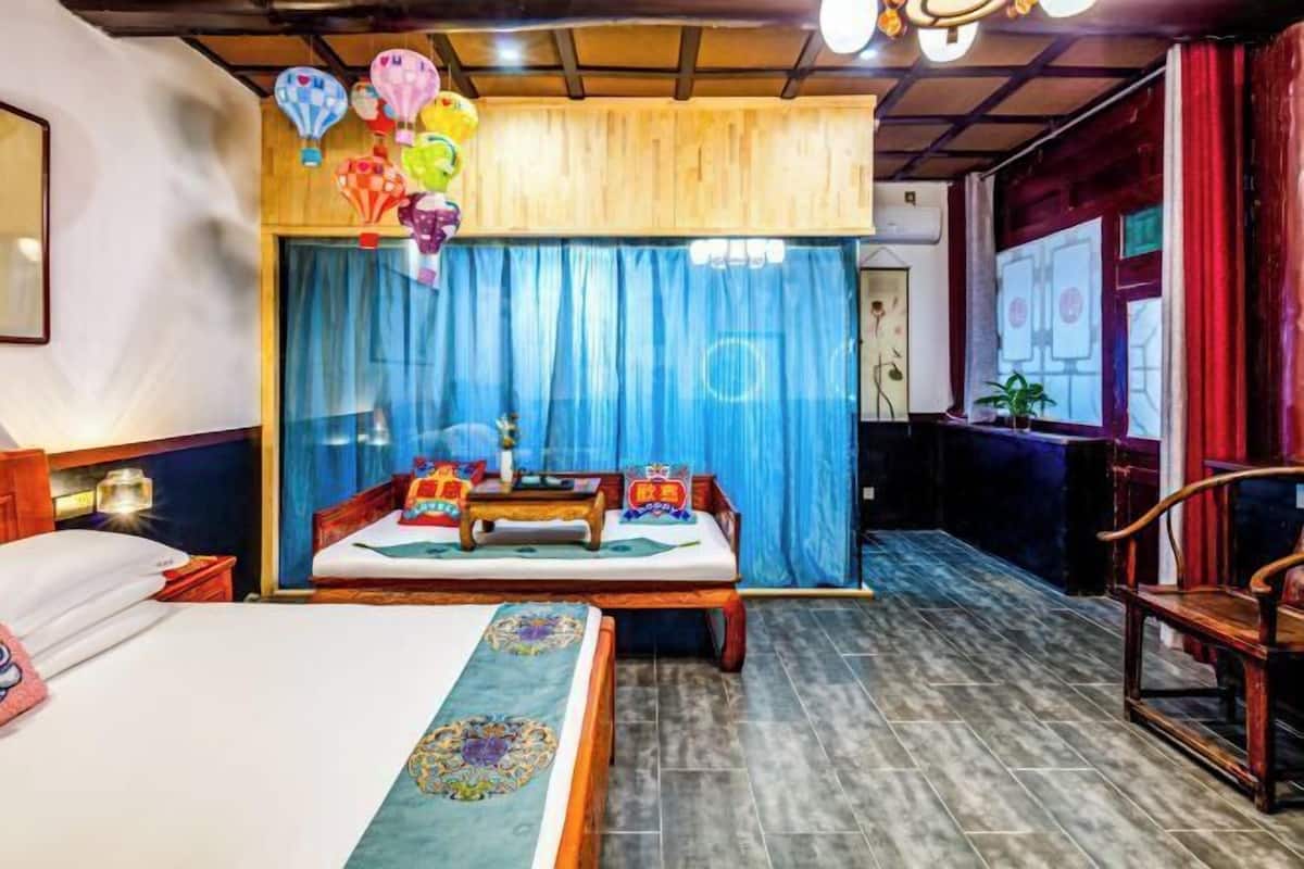 Ein Hotelzimmer mit Bett, Sofa, kleinem Tisch und Stuhl. Vorhanden sind ein Vorhang, eine Pflanze und eine Wand mit einem Gemälde.