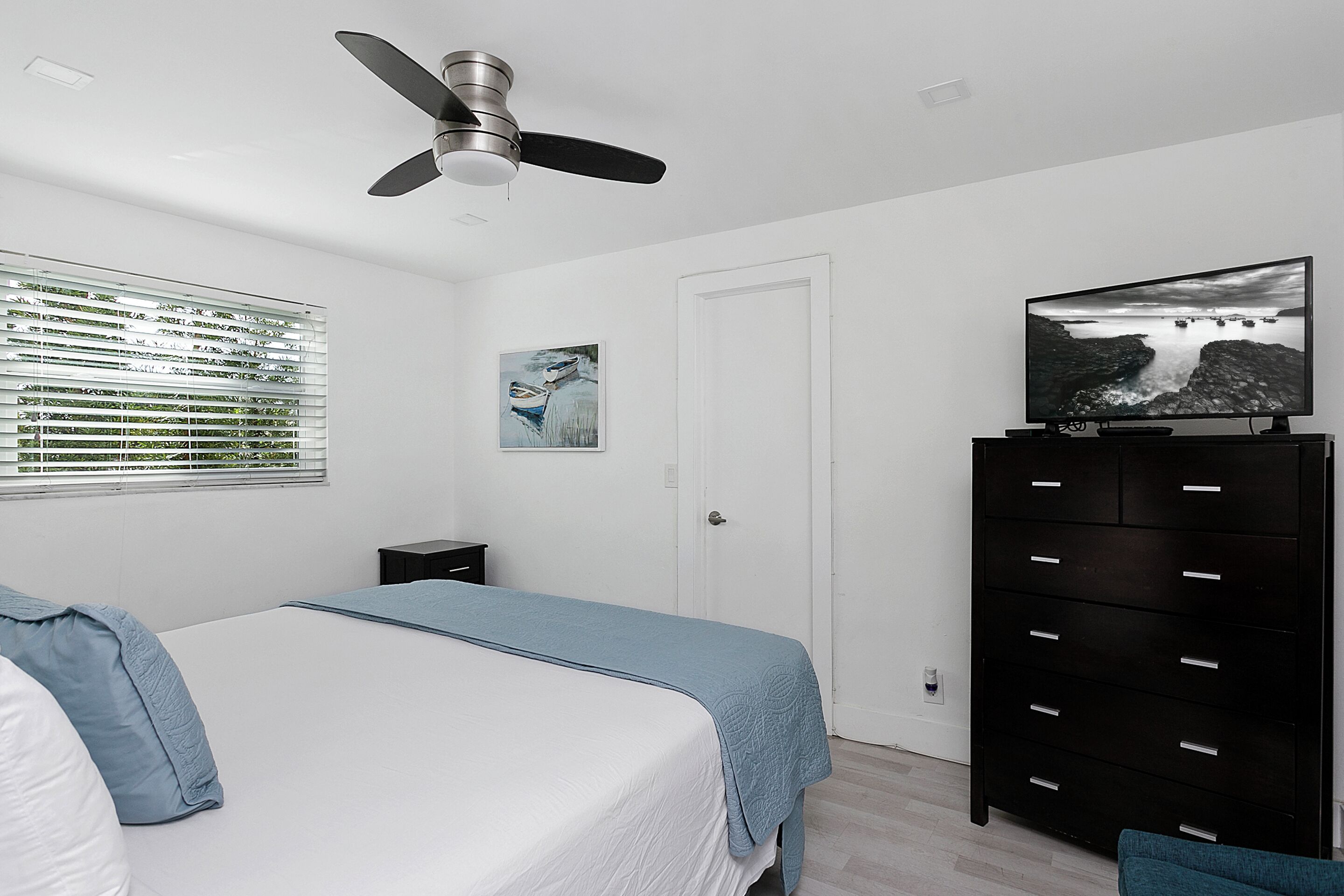 Un dormitorio con una cama, un ventilador de techo, un televisor sobre un aparador, una ventana con persianas y un cuadro en la pared.