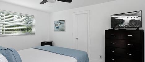 Un dormitorio con una cama, un ventilador de techo, un televisor sobre un aparador, una ventana con persianas y un cuadro en la pared.