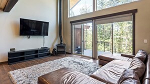Ein großzügiger Wohnraum mit einer breiten Ledersofa, einem Flachbildfernseher, einem Kamin und einer Glastür, die auf einen Balkon mit Blick auf Bäume führt.