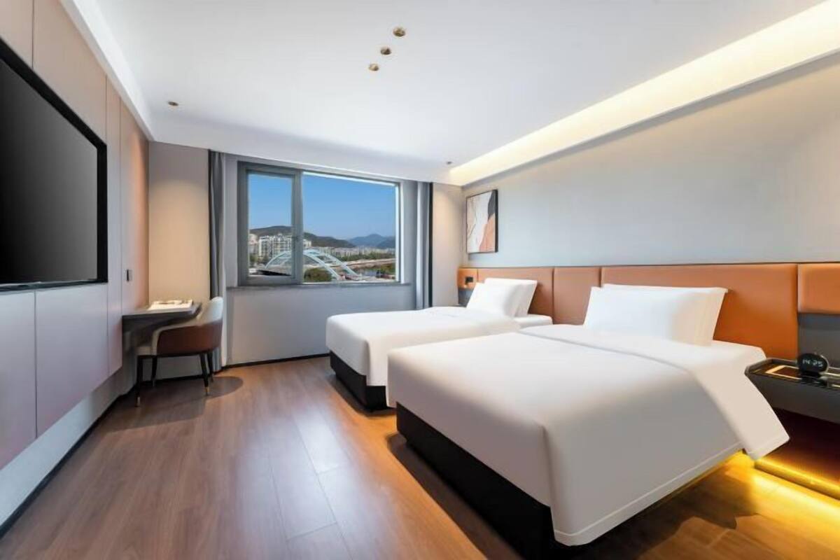 Habitación de hotel moderna con dos camas, un escritorio, una silla y un amplio ventanal con vista a la ciudad.