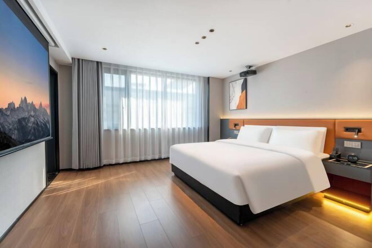 Habitación de hotel moderna con una cama grande, vistas a montañas a través de la ventana y un mural de un paisaje urbano en la pared.