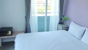 Une chambre d’hôtel avec un grand lit, une table de chevet avec une radio, une petite table de nuit avec un vase, et une vue sur un immeuble par la fenêtre.