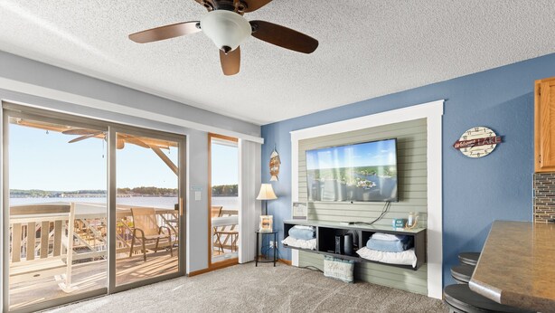 A spacious living area with a large sliding glass door leading to a deck, a ceiling fan, a television mounted on the wall, and a small bar area. - Golden Reef! NEW pool! State park views! GR102 (Osage Beach)
