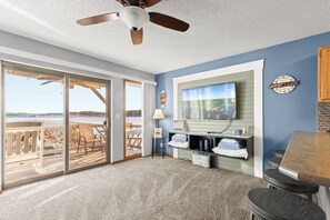 A spacious living area with a large sliding glass door leading to a deck, a ceiling fan, a television mounted on the wall, and a small bar area. - Golden Reef! NEW pool! State park views! GR102 (Osage Beach)