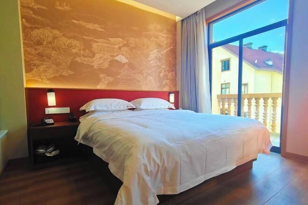 A hotel room with a large bed, a nightstand with a lamp, a wall with a mural, and a balcony with a railing.