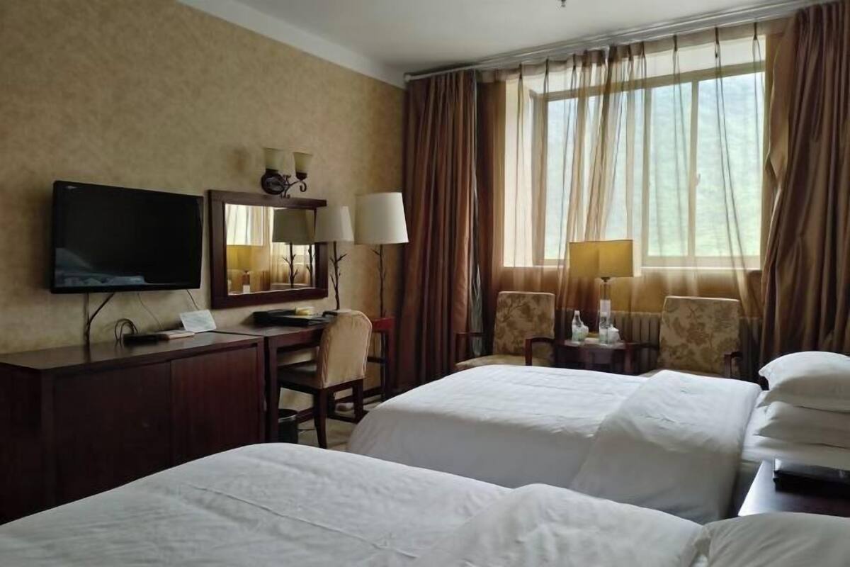 A hotel room with two beds, a desk, a television, and a large window with curtains.