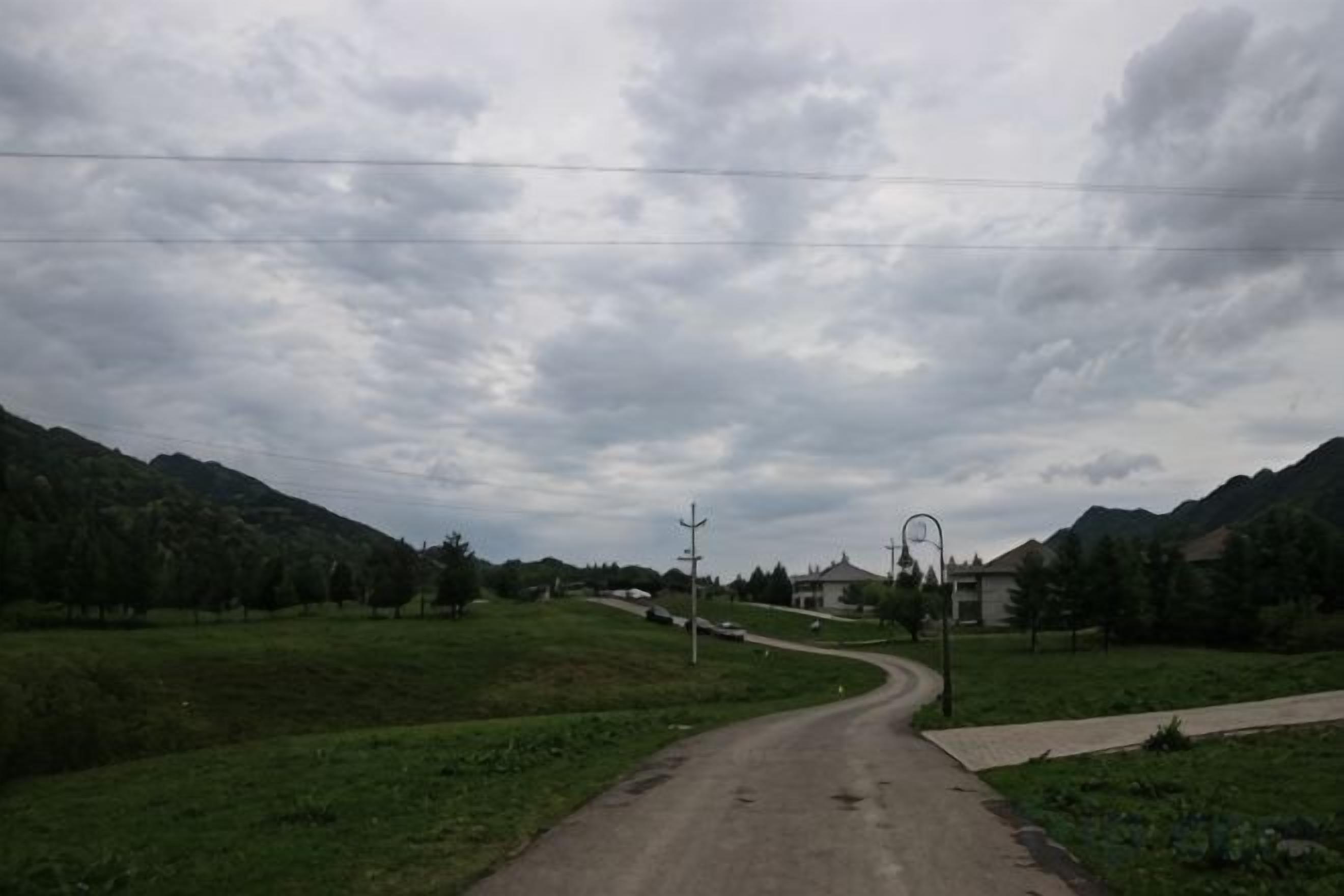 A rural road curving through a green landscape with houses and power lines in the distance.