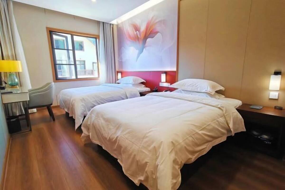 A hotel room with two beds, a desk with a lamp, a large window, and a wall painting.