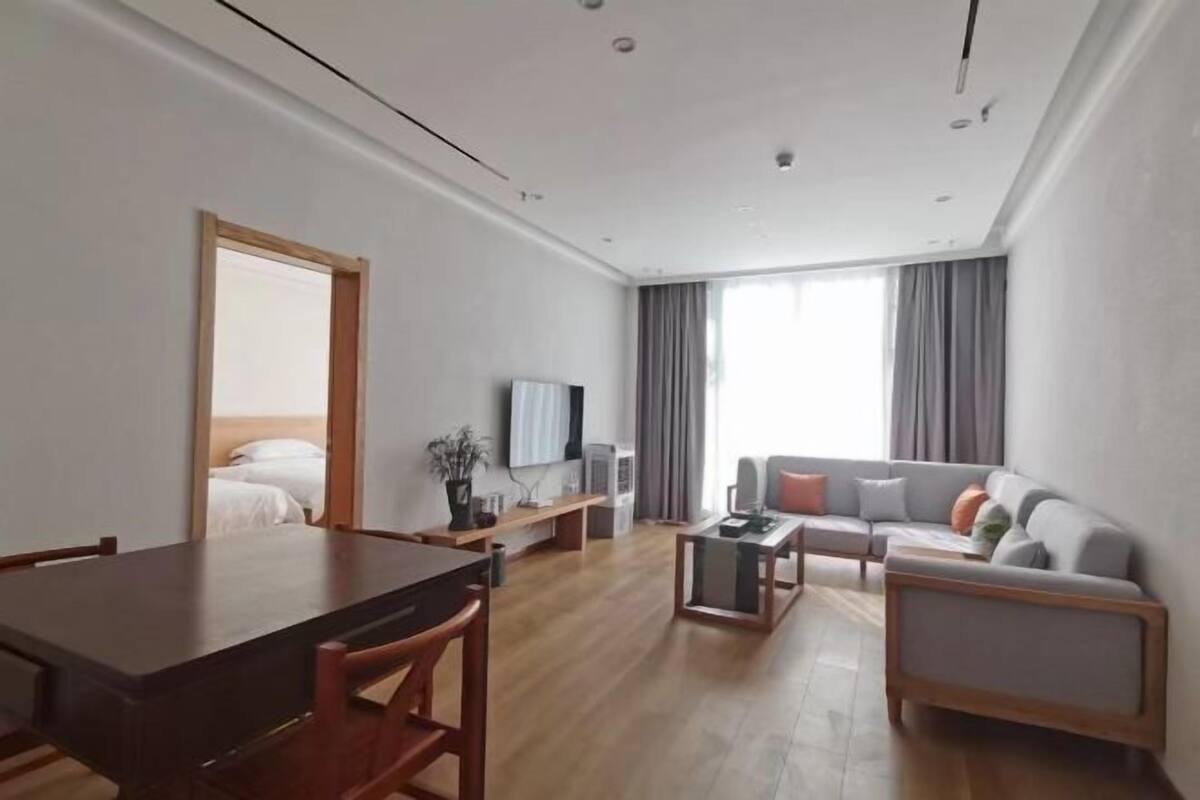 A modern hotel room with a wooden dining table, a sofa, a TV, and a large window with curtains.