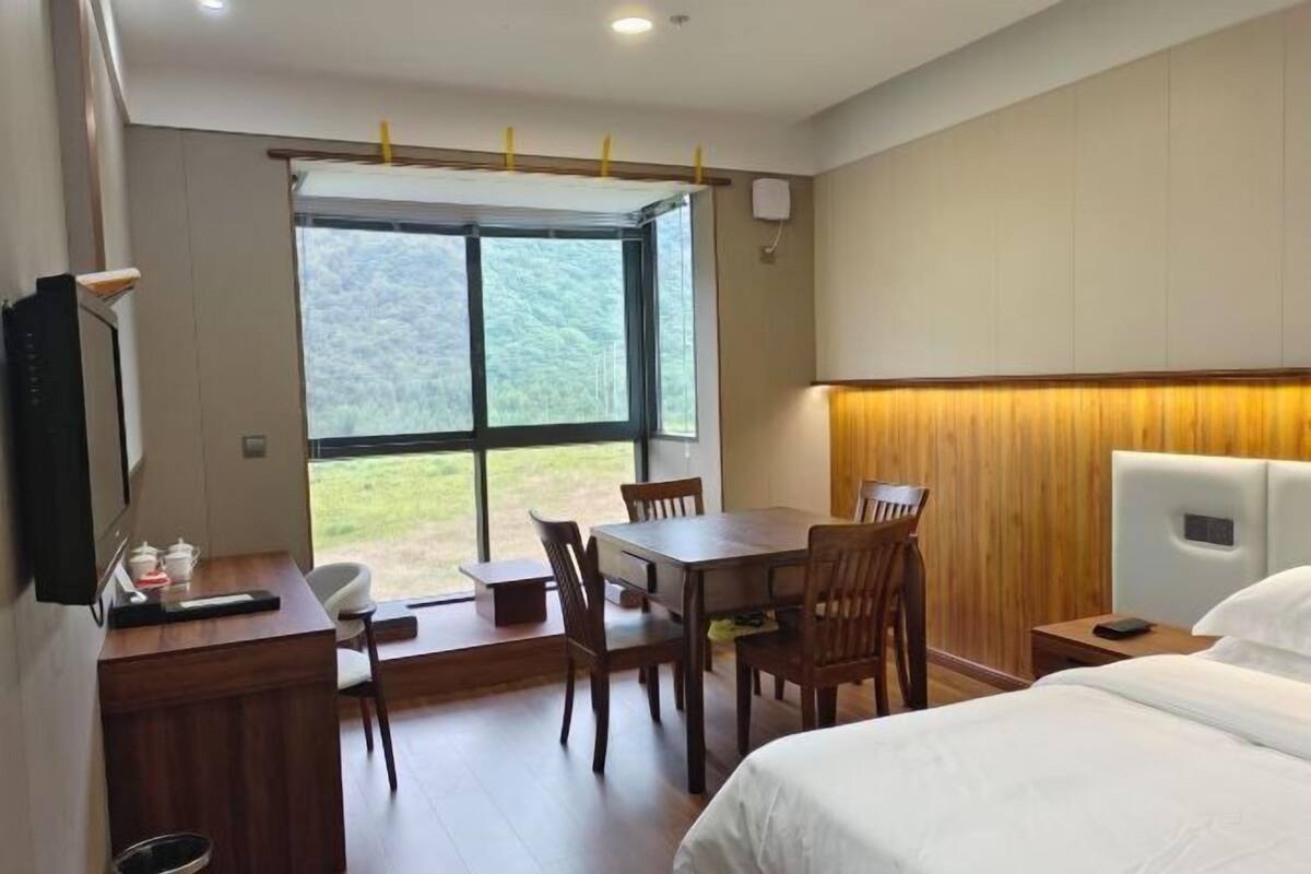 A hotel room with a bed, a desk with a chair, a table with chairs, and a large window with a view of greenery.