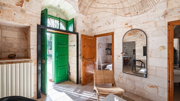 Une pièce aux murs en briques apparentes, avec une porte verte, une porte en bois, une chaise en osier, une table sur laquelle se trouve une tasse et un miroir accroché au mur.