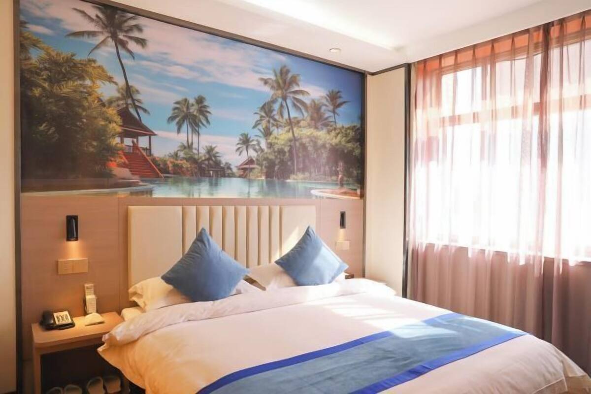 Une chambre d’hôtel avec une grande fresque murale sur le thème des tropiques, un lit avec une literie blanche et bleue, et une table de chevet avec un téléphone et une lampe.
