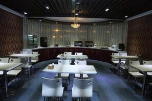 Un salón de conferencias con disposición curva, mesas y sillas blancas, una zona de bar central y una araña.