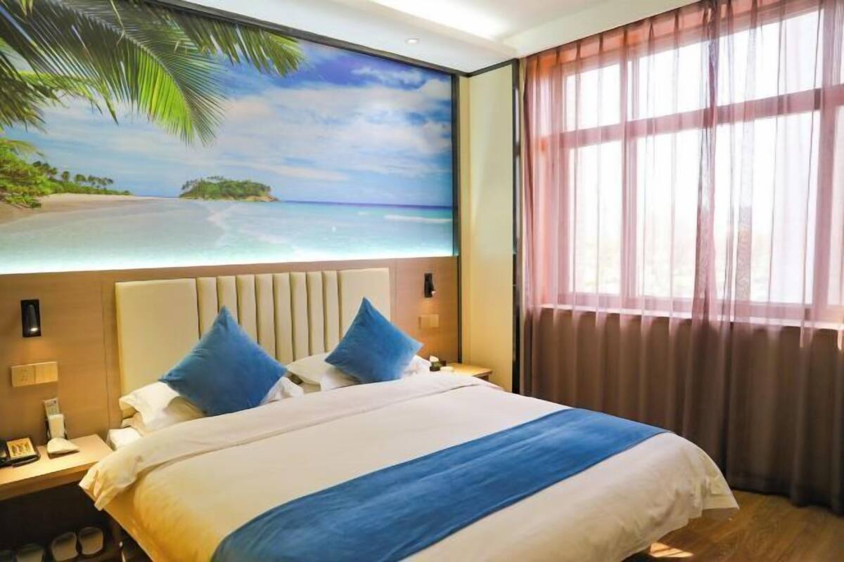 Une chambre d’hôtel avec un lit, une table de chevet, une fresque murale représentant une plage et une fenêtre avec des rideaux.