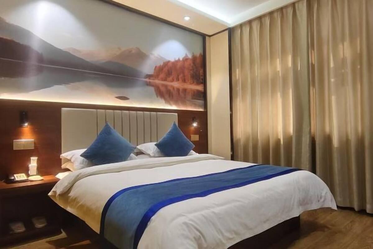 Une chambre d’hôtel avec un grand lit, une tête de lit ornée d’un tableau, une table de chevet avec un téléphone et une fenêtre avec des rideaux.