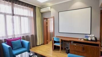 Een moderne hotelkamer met een groot raam, een zithoek met blauwe banken, een houten bureau en een groot scherm.