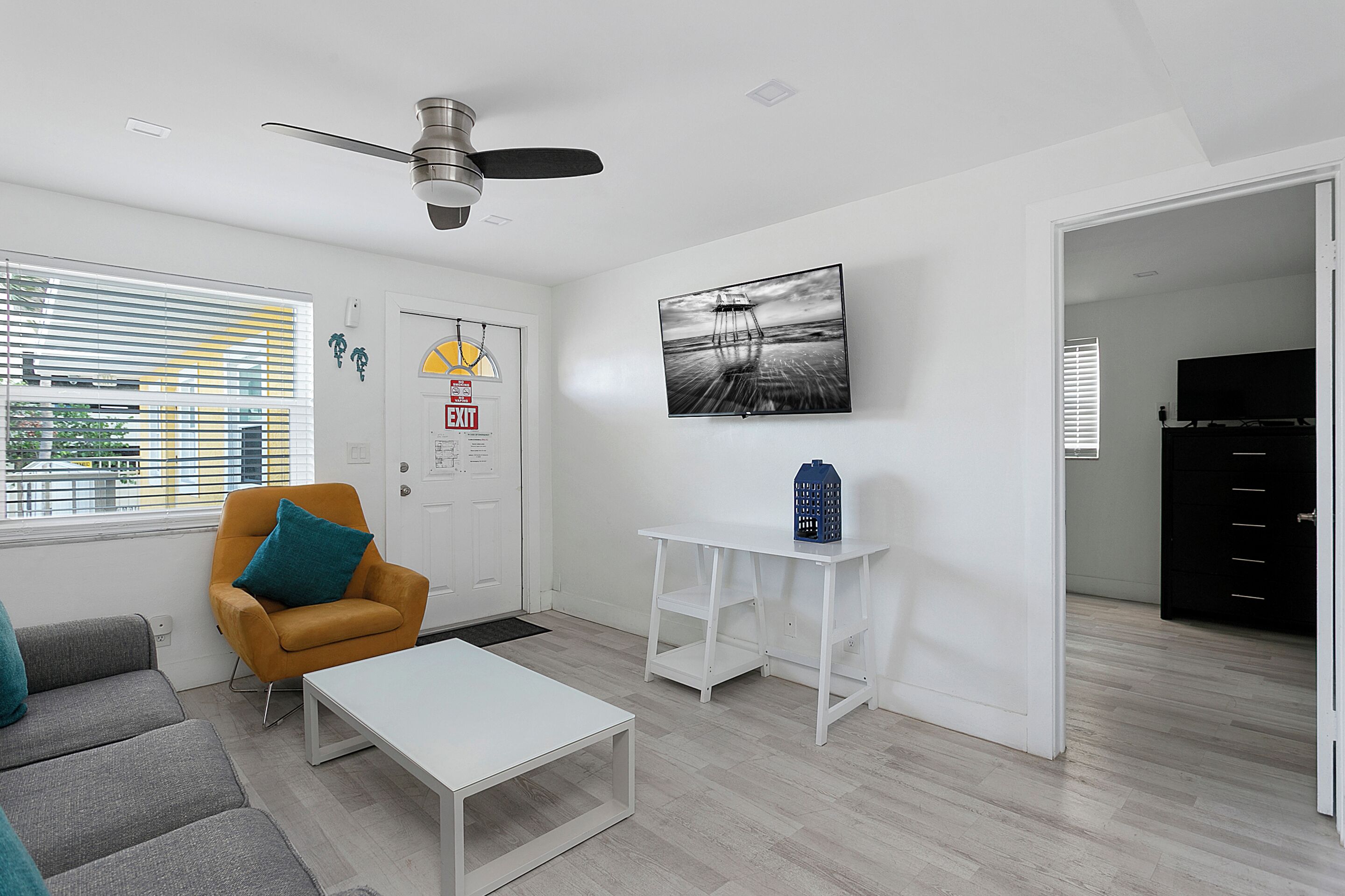 A modern living room with a white ceiling fan, a sofa with teal cushions, a white coffee table, and a painting of a bridge.