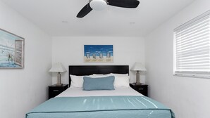 A neatly arranged bedroom with a bed, two bedside tables with lamps, a ceiling fan, a beach-themed painting, and a window with blinds. - ULTRA joyful apartment great for family (Hollywood)