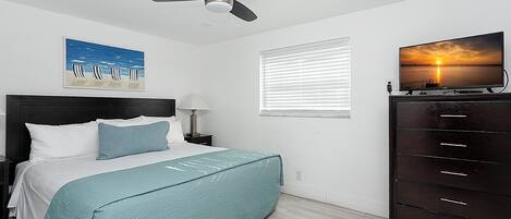 Un dormitorio con cama, ventilador de techo, televisor en una cómoda y un cuadro de una playa.