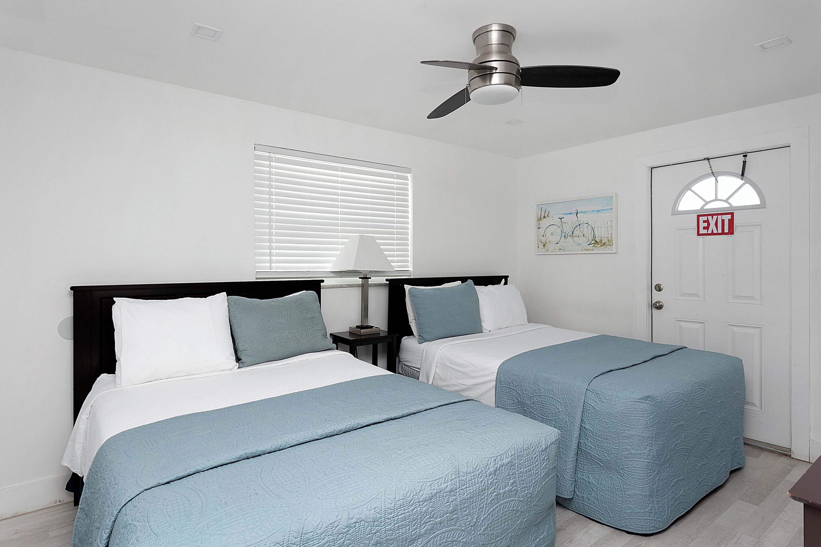 A bedroom with two beds, a ceiling fan, a window with blinds, a nightstand with a lamp, and an exit sign.