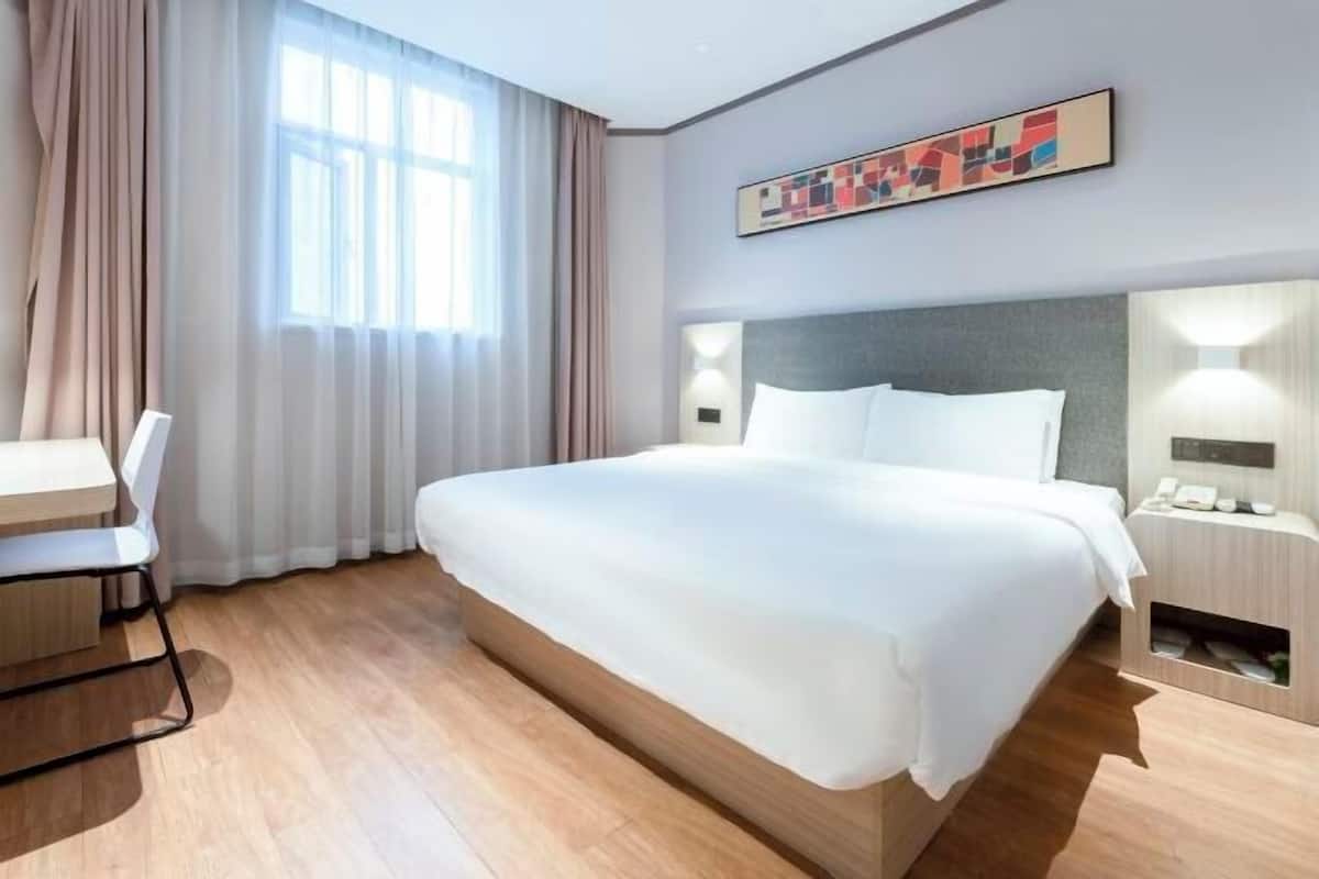 A hotel room with a large bed, a desk, a chair, a bedside table, and a window with curtains.