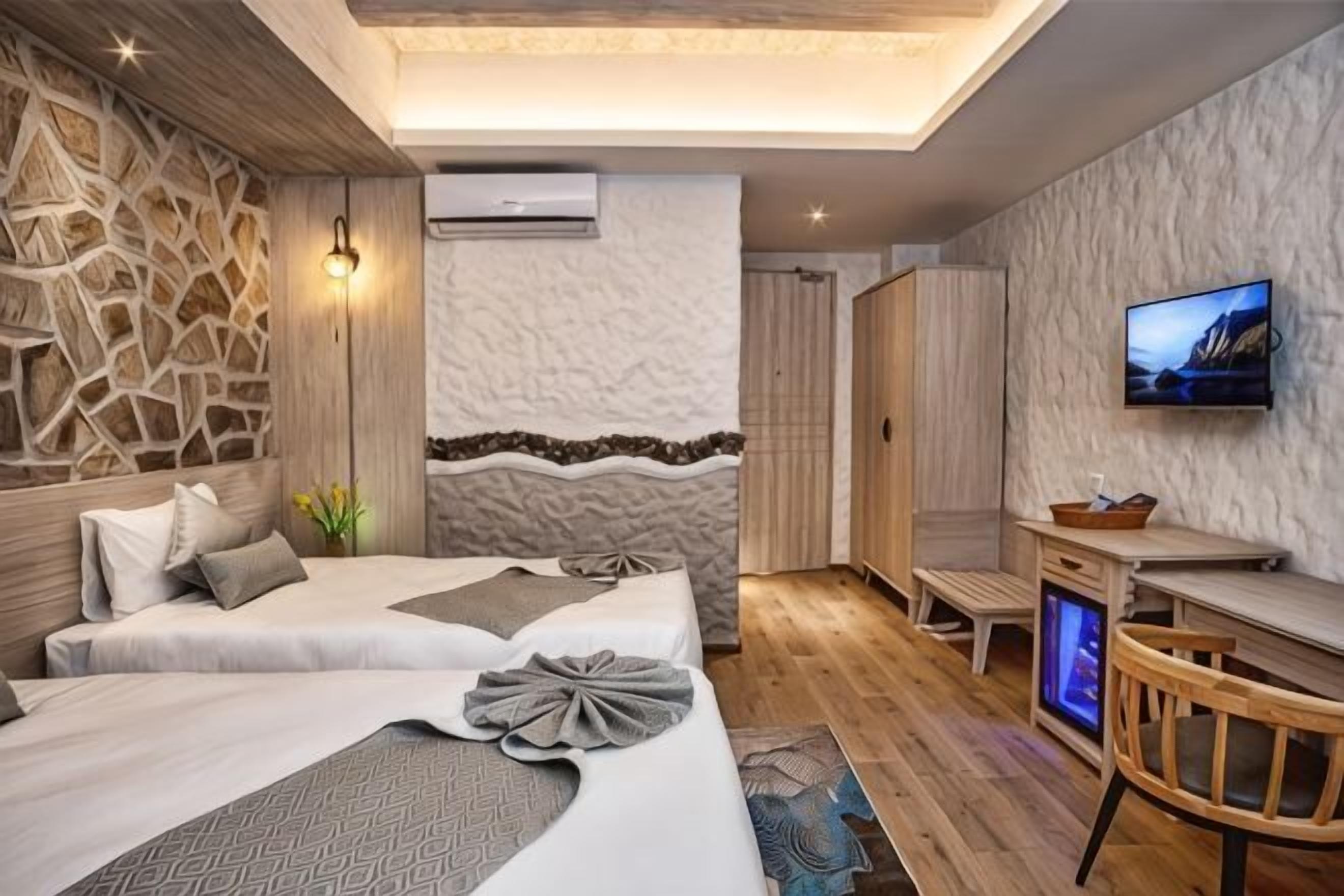Quarto de hotel moderno com parede de pedra, televisão de tela plana, cama com colcha estampada e uma pequena mesa de madeira.