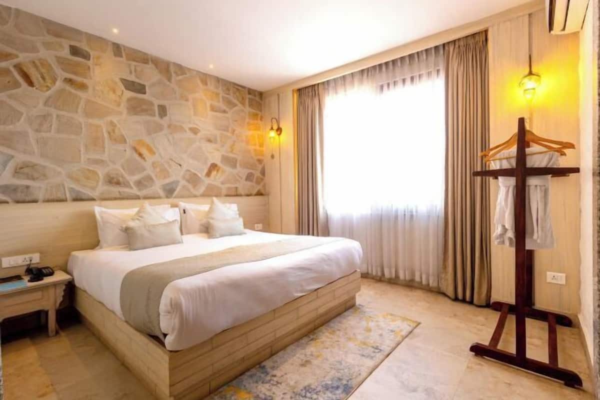 Une chambre avec un mur en pierre, un lit avec une literie blanche, une table de chevet avec un téléphone, un cintre en bois et une fenêtre avec des rideaux.