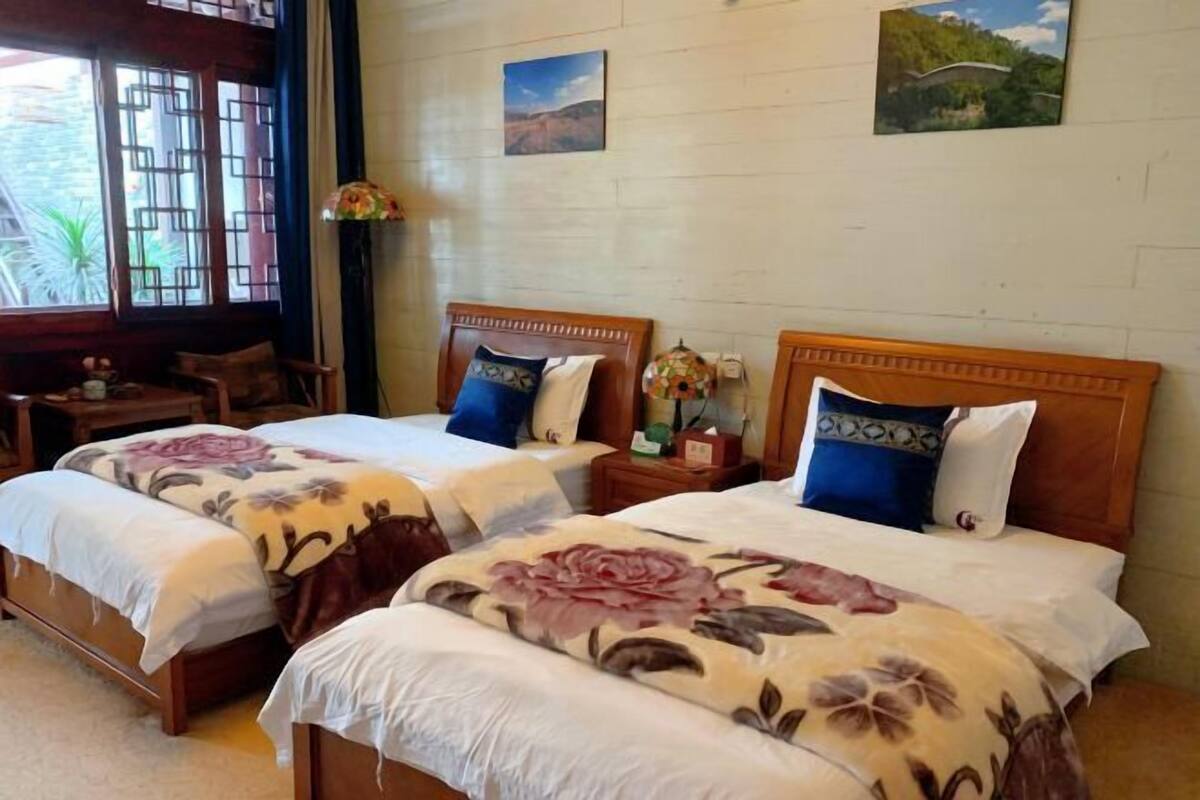 A hotel room with two beds, wooden headboards, floral bedspreads, a small table, and a window with a view of greenery.