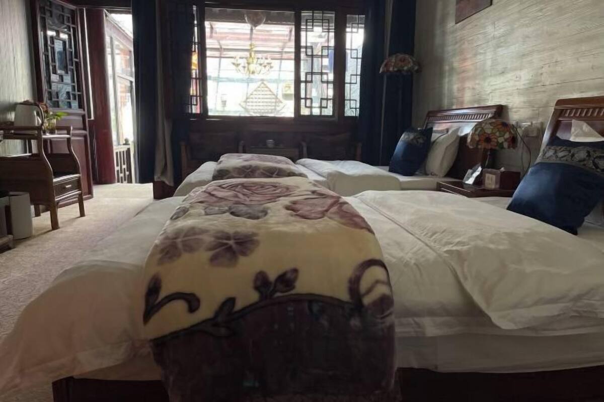A hotel room with three beds, a wooden desk, a chandelier, and a view through a window with geometric patterns.