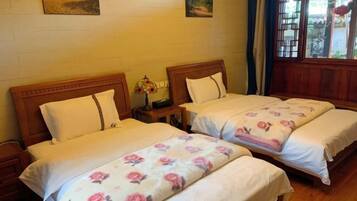 A room with two beds, each with white bedding and floral patterns. There are wooden headboards and nightstands with lamps. The room has a wooden floor and a window with a view of the outside.