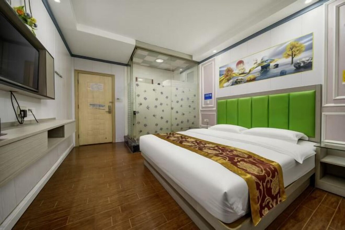 A hotel room with a bed, a television, a green headboard, and a painting on the wall.