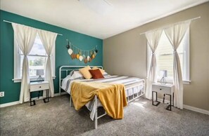 Un dormitorio con paredes azul verdoso, una cama con manta amarilla, dos mesitas de noche y cortinas en las ventanas.